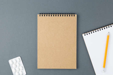 Open notebook with clear page with yellow pencil and tablet blister on grey background. Design...