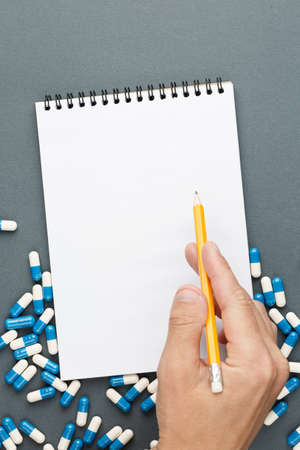 Open spiral notebook with clear page male hands with pencil and pills on grey background. Design business, medicine supplies or medical concept. Ready for adding or mock up. Top view. Vertical photo.の写真素材