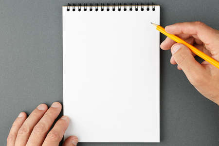 Open spiral notebook with clear page and male hands with yellow pencil on grey background. Design...