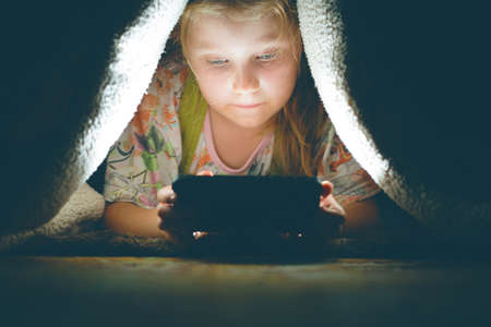 girl plays games on a smartphone at night under a blanket on the floor, the child rewrites with...