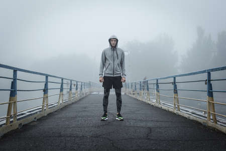 Athlete in sportswear stands in the middle of a bridge on a foggy morning. The beginning of the morning workout on a natural background.の写真素材