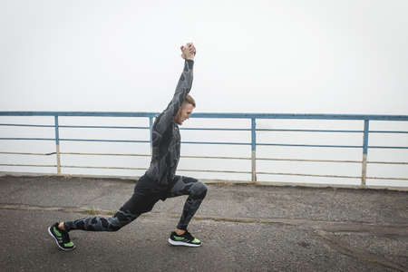 Athletic man in compression clothing stretches on a bridge before running against a background of gray morning fog.の写真素材