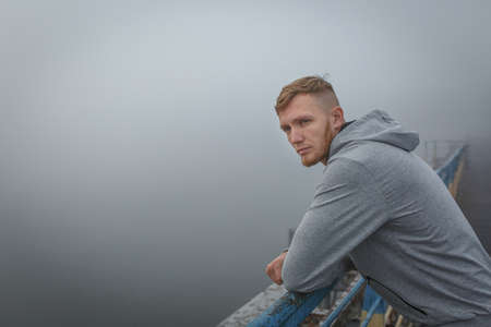 Handsome man in a gray hoody leans on the iron handrails of a bridge against the backdrop of a foggy...