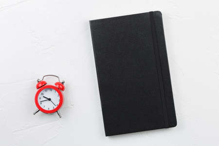 Black leather notepad with red alarm clock on white textured table. Notebook goals plans dreams,...