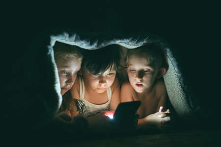 children play games on a smartphone at night under a blanket on the floor, the child rewrites with friends on social networksの写真素材
