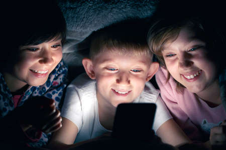 Joyful children under the blanket at night in theirs bed communicates, smiling and having fun on Internet. Child gadget addiction and insomnia.の写真素材