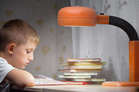 Cute little boy doing homework at the table reading textbooks in the evening under the light of the lamp distance education and learningの写真素材