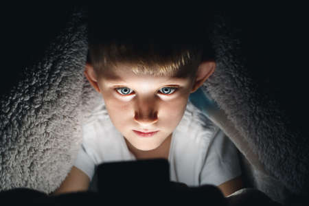 Boy with a serious face under the blanket at night in his bed communicates on Internet. Child gadget addiction and insomnia.の写真素材