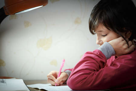Little pretty girl writes with a pen in a notebook does her homework in the evening at home by the...