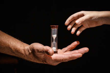 Adalt hand passes the glass hourglass to the child Concept of the generation change On a black isolated background.の写真素材