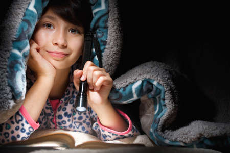Nice girl lies under a blanket at night reading a book with a flashlight.