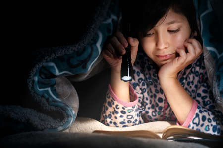 Nice girl lies under a blanket at night reading a book with a flashlight.の写真素材