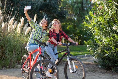 Two pretty young caucasian girls making selfie photo with smartphone. Best friends enjoying a day on bikes. Sunny summer evening.の写真素材