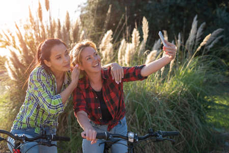 Two pretty young caucasian girls making selfie photo with smartphone. Best friends enjoying a day on bikes. Sunny summer evening.の写真素材