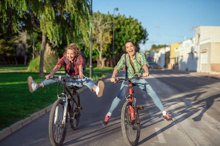 Two pretty young caucasian girls having fun on bicycles along the street. Best friends enjoying a day on bikes. Sunny summer evening.の写真素材