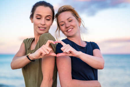 leisure and friendship concept - two happy smiling teenage girls or best friends at seaside making hand heart gesture. selective focus.の写真素材