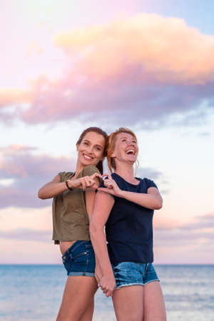 leisure and friendship concept - two happy smiling teenage girls or best friends at seaside making hand heart gesture at sunset.の写真素材