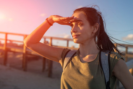 Fashionable slender young brunette girl with backpack posing on a background wooden walkways and sand beach at sunset.の写真素材