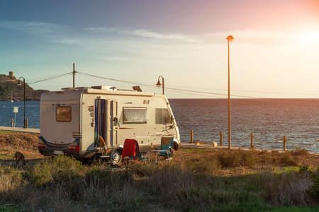 Trailer motor home and pet dogs on the grassy part of the beach at sunset. Leisure mobile camping home for tourists overlooking the blue sea and cape. Adventure relaxing travel on caravan van.の写真素材