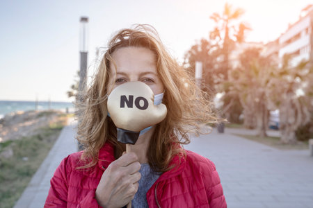 Young woman in protective sterile medical mask on her face, on street background, holding small boxing glove with No sign. America president election, pandemic virus concept.の写真素材