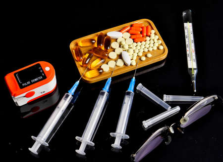 Pulse oximeter, syringes, tablets on a plate on a mirrored black background. Close up.