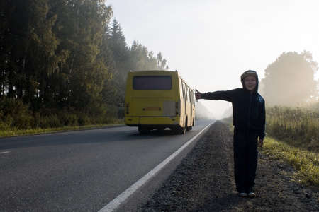 A boy in a dark hooded sweatshirt hitchhiking on a forest road in a fogの写真素材