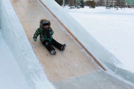 A boy in a green jacket and striped cap slides on an ice slideの写真素材
