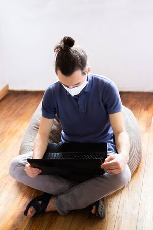 A white man using laptop at home during COVID-19 quarantine. Caucasian man wearing medical mask and working remotely at home and wearing face mask for virus prevention. Coronavirus concept.の写真素材
