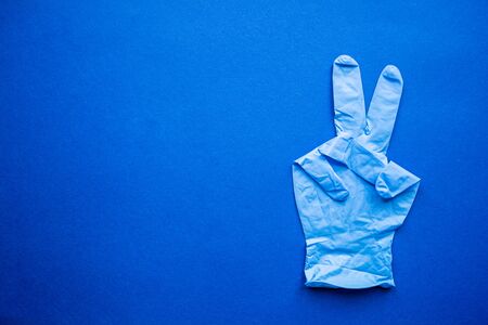 Medical blue glove shows victory sign on blue background. Health protection and defeating the coronavirus pandemic concept.の写真素材