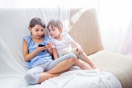 Children at home sitting on sofa, playing with tablet. Cute siblings brother and sister have a good time at home. Kids play a game on a cell phone. Stay at home during quarantine COVID-19 conceptの写真素材