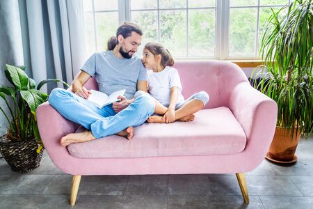 Fathers day concept. Amazing father and cute little girl reading a book, smiling and enjoying at home. Happy parenthood. Father and daughter spending time together.の写真素材