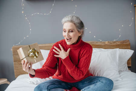 A beautiful emotional senior woman with gift at home on Christmas lights background