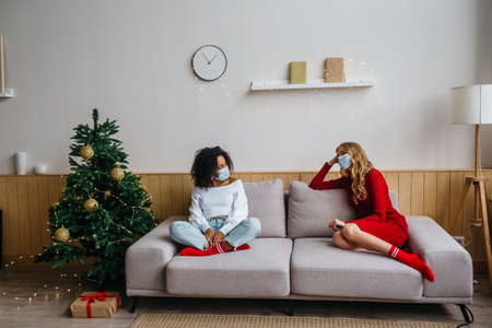 Social distance while celebrating Christmas and New Years 2021 concept. Two girls in protective masks sitting on the couch 1 m from each other.の写真素材