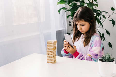 Focused little girl recording video or take photo of board game with wooden blocks. Little blogger at homeの写真素材