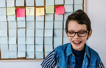 Portrait of cheerful autism boy with glasses in blue shirt front a board with stickers and the note Autism. Happy autistic boy laughing.の写真素材
