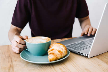 Close up young man hands on the wooden table with laptop, cappuccino and croissant. Freelance and work from a cafe conceptの写真素材