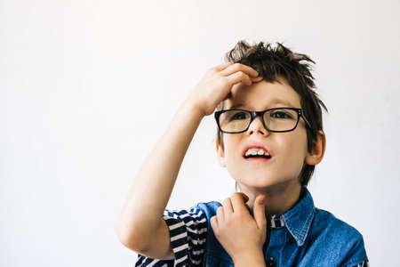 Autism boy with glasses in blue shirt on a white wall backdrop holds his head with his hands, thinks and tries to remember. Autism awareness and pain conceptの写真素材