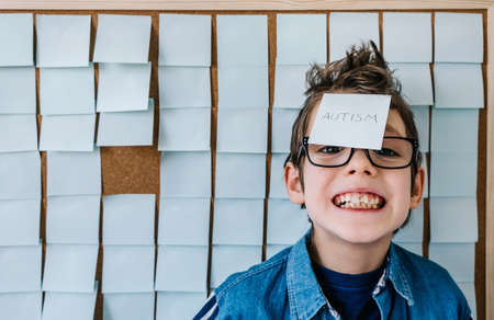 Autism awareness funny concept. Portrait of cheerful autism boy with glasses in blue shirt front a board with stickers and the note Autism.の写真素材