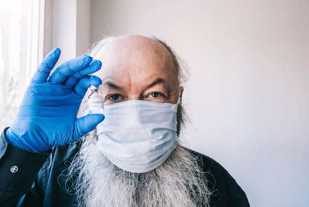 vaccination for the elderly. Portrait of an bearded senior man wearing a medical mask and holding an ampoule with a vaccine.の写真素材