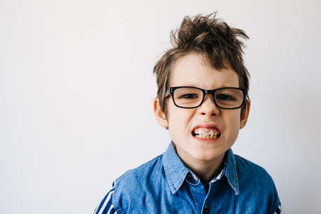 Portrait of autism boy with glasses in blue shirt on a white wall backdrop grimaces. Autism awareness and pain conceptの写真素材