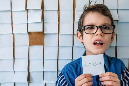 Autism awareness concept. Portrait of cheerful autism boy with glasses front a board with stickers and holds the note Autism.の写真素材