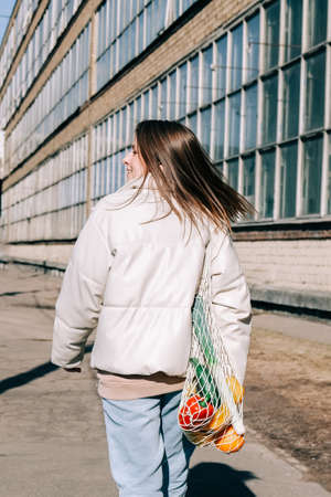Reusable recyclable lifestyle concept. Young woman A young woman walks through the city and holds a reusable mesh shopping bag with fruits and vegetables.の写真素材
