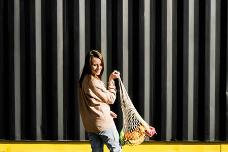 Trendy colors zero waste concept. Cheerful young woman smiling and holding reusable mesh shopping bag with fruits and vegetables.の写真素材
