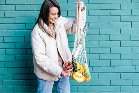 Zero waste eco living concept. Happy woman smiling and holding reusable mesh shopping bag with fruits and vegetables.の写真素材