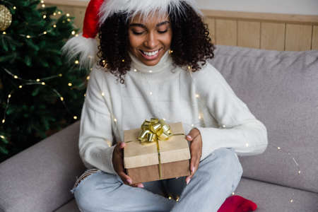 Close up portrait of cheerful smiling african american millennial woman in santa hat opens a christmas gift. Festive lifestyle conceptの写真素材