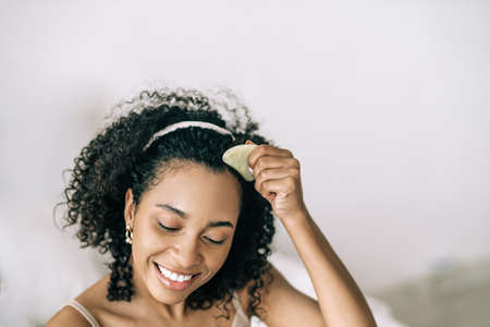 Lifestyle portrait of smiling beautiful african american woman using facial gua sha jade. Natural self skin care and anti-aging conceptの写真素材