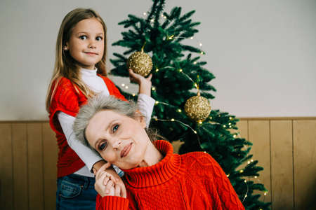 Merry Christmas family concept. Elegant gray-haired grandma and her cute grand daughter girl durind Cristmas getting ready. Granny and child near tree indoors.の写真素材