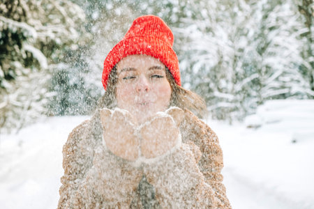 Young woman in trendy winter clothes and red hat blowing snowflakes, which holds in her hands in...