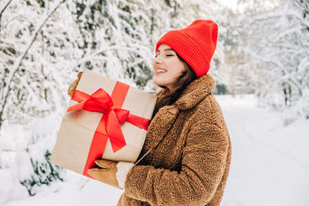 Christmas gifts and shopping concept. Close up portrait of young woman wearing red hat holds big red box with red ribbon in her hands outside in the winter snowy forest.の写真素材