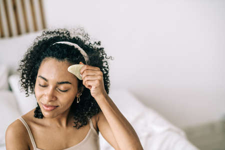 Natural beauty care and anti-aging concept. Lifestyle portrait of beautiful woman using stone facial gua sha jade in bedroomの写真素材